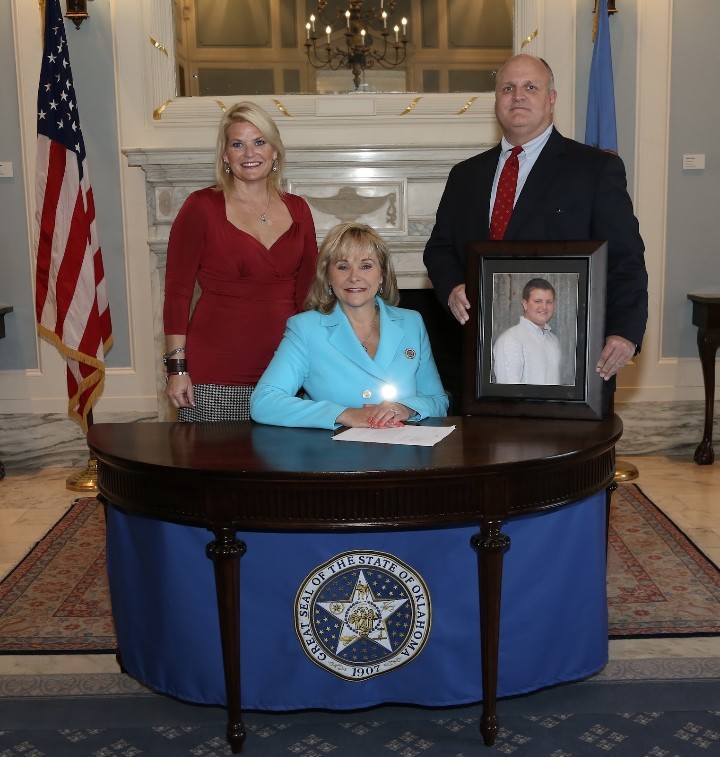 Governor Mary Fallin seated at a desk bearing the Great Seal of Oklahoma, signing Senate Bill 239 into law on June 10, 2015. Chase Morris's father stands to her right, holding a framed portrait of Chase.